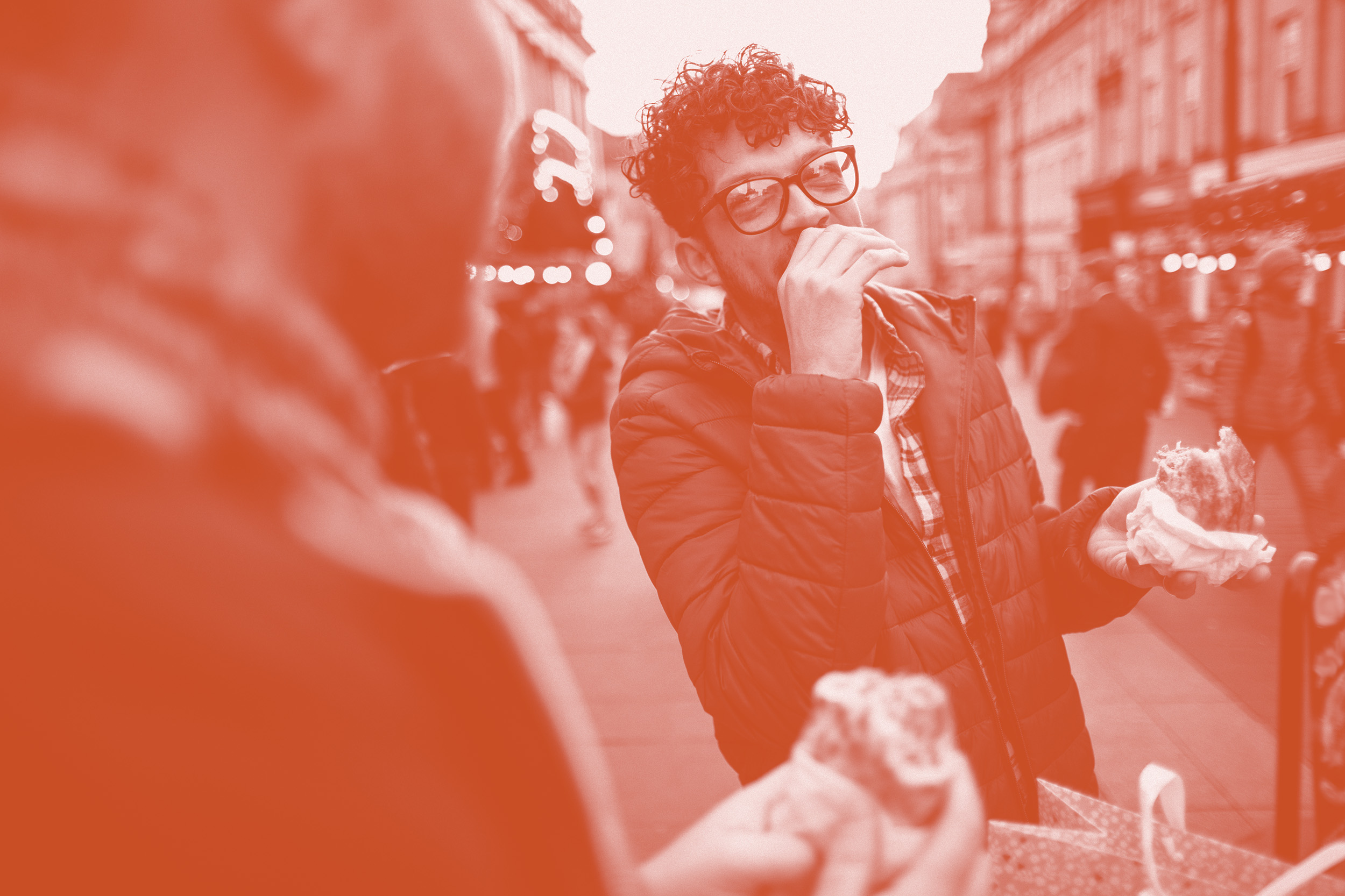 Man on a high street eating food while appearing to be amused by something someone else has done or said
